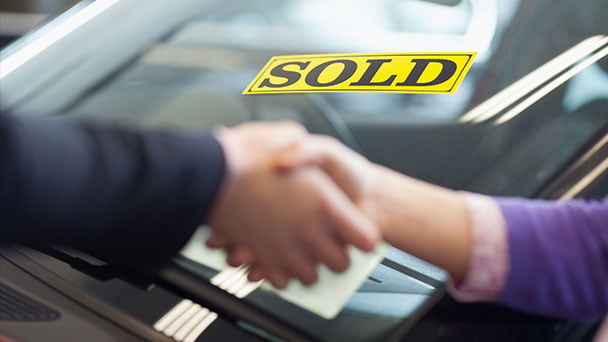 Buyer and seller shaking hands in front of Sold sign on vehicle that has just been bought.