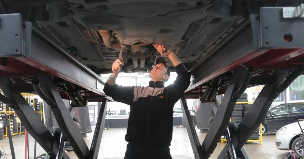Man in black jacket standing under a car doing some work.