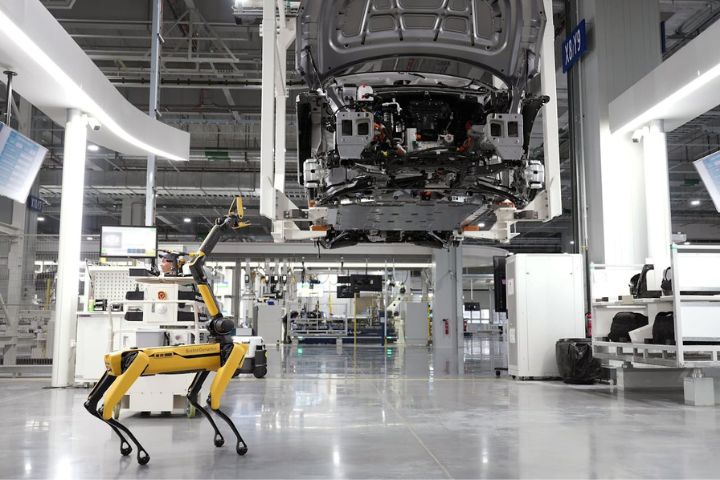 A yellow quadruped robot inspects a partially assembled car suspended in a high-tech automotive manufacturing facility.