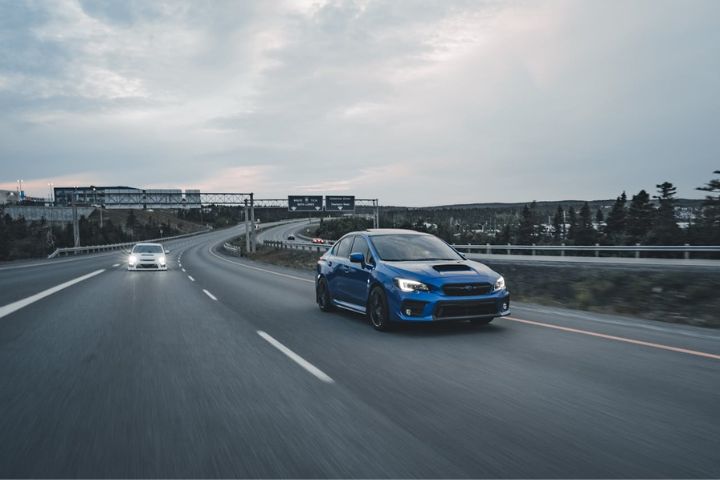 A blue Subaru WRX leads on an open highway at dusk, followed by another car with headlights on, against a backdrop of trees and distant road signs.