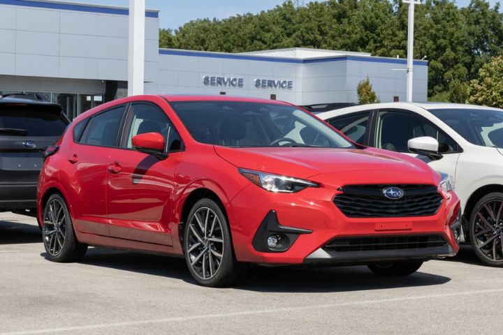 A red Subaru Impreza hatchback is parked in a dealership lot, positioned in front of a service center building on a sunny day.